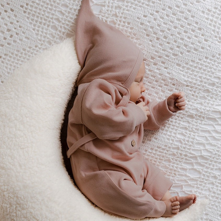 Een vredig kindje gekleed in een gezellig bruin Boxpakje Blush gemaakt van hoogwaardig katoen van Yumi Baby, met een bijpassend hoedje, opgerold en duttend op een zacht wit halvemaanvormig kussen.