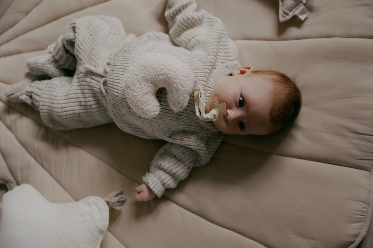 Gezellig en comfortabel, een kleintje gekleed in een Yumi Baby Knitted broek Salt & Pepper-outfit doet een vredig dutje op een zachte ondergrond, met een pluchen sterrenkussen en redelijke slofjes in de buurt.