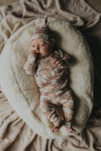 Een serene pasgeboren baby gekleed in een Yumi Baby Broekje Boho Jungle onesie gemaakt van OEKO-TEX gecertificeerd katoen en een bijpassend mutsje, vredig slapend in een knus rieten mandje gevoerd met een zachte, crèmekleurige deken.