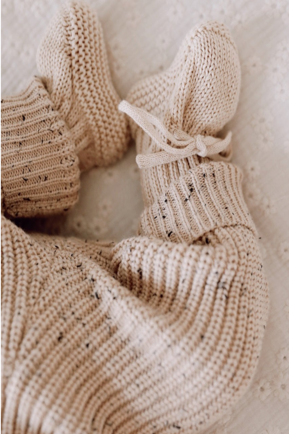 Een paar knusse, 100% katoenen Yumi Baby handgebreide beige slofjes, waarschijnlijk voor een baby van 0-12 maanden, vastgebonden met delicate strikjes, die zachtjes tegen elkaar rusten.