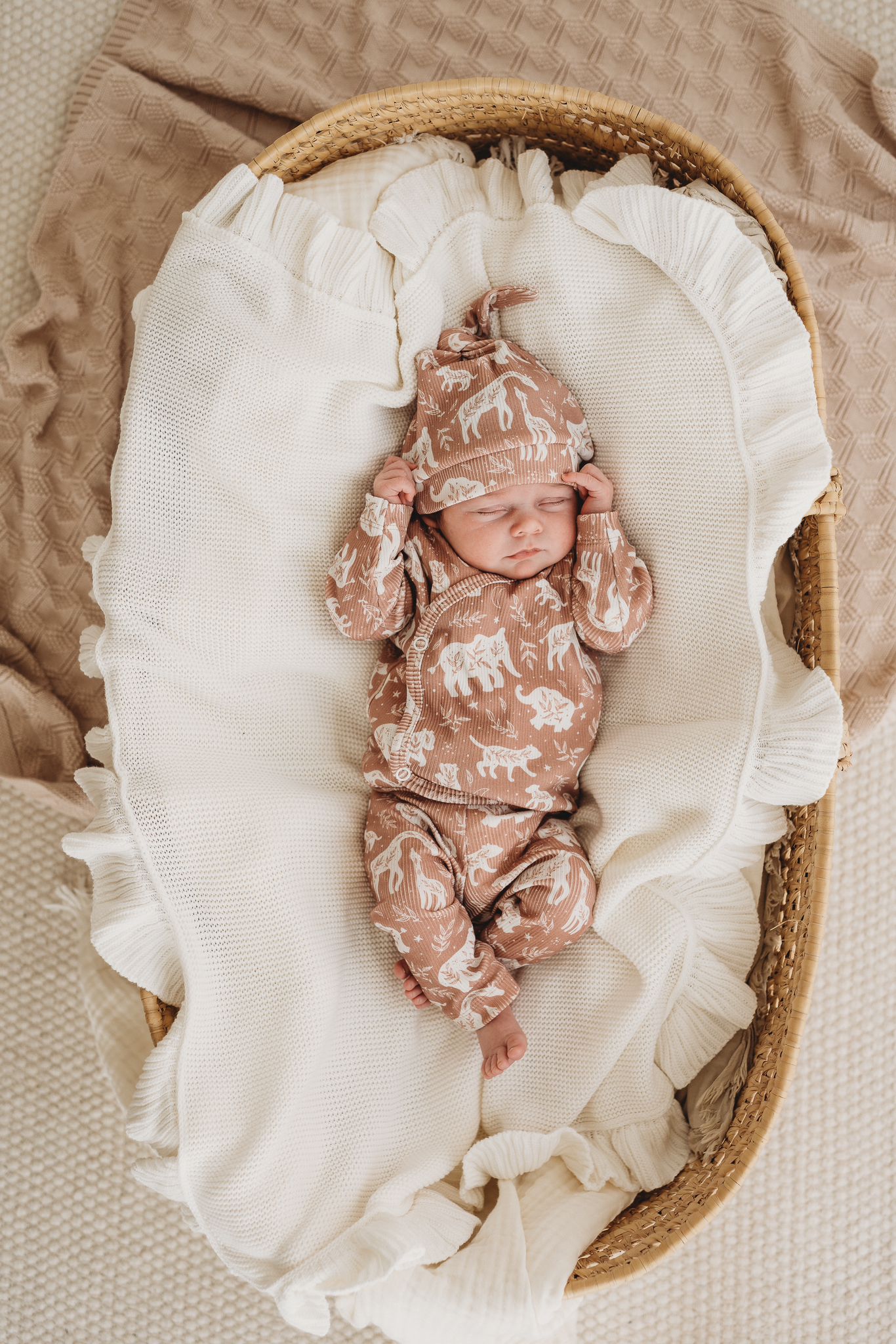 Een vredige pasgeboren baby gekleed in een rompertje met patroon en bijpassende Muts Boho Jungle, heerlijk slapend in een knus mandje omringd door zachte, crèmekleurige dekens.