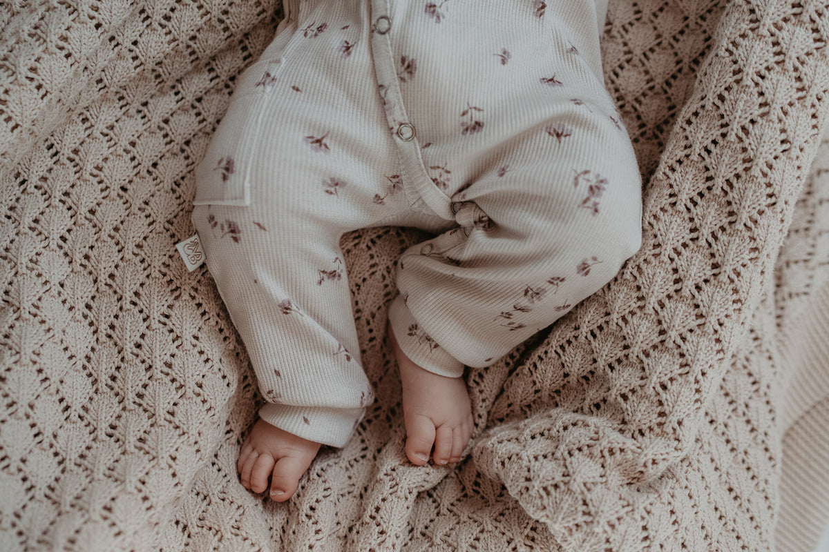 Een vredig slapende pasgeboren baby, gekleed in een witte Yumi Baby Oeko Tex katoenen onesie met delicate paarse bloemmotieven, rustend op een crèmekleurige deken met een gegolfde rand.