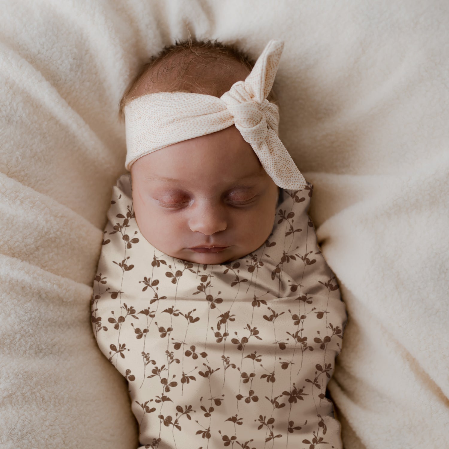 Een serene pasgeboren baby ingebakerd in een gebloemde Yumi Baby Inbakerdoek Brown Leafs omslagdoek, zachtjes slapend op een zachte, crèmekleurige deken met een schattige strikhoofdband die haar hoofd siert.
