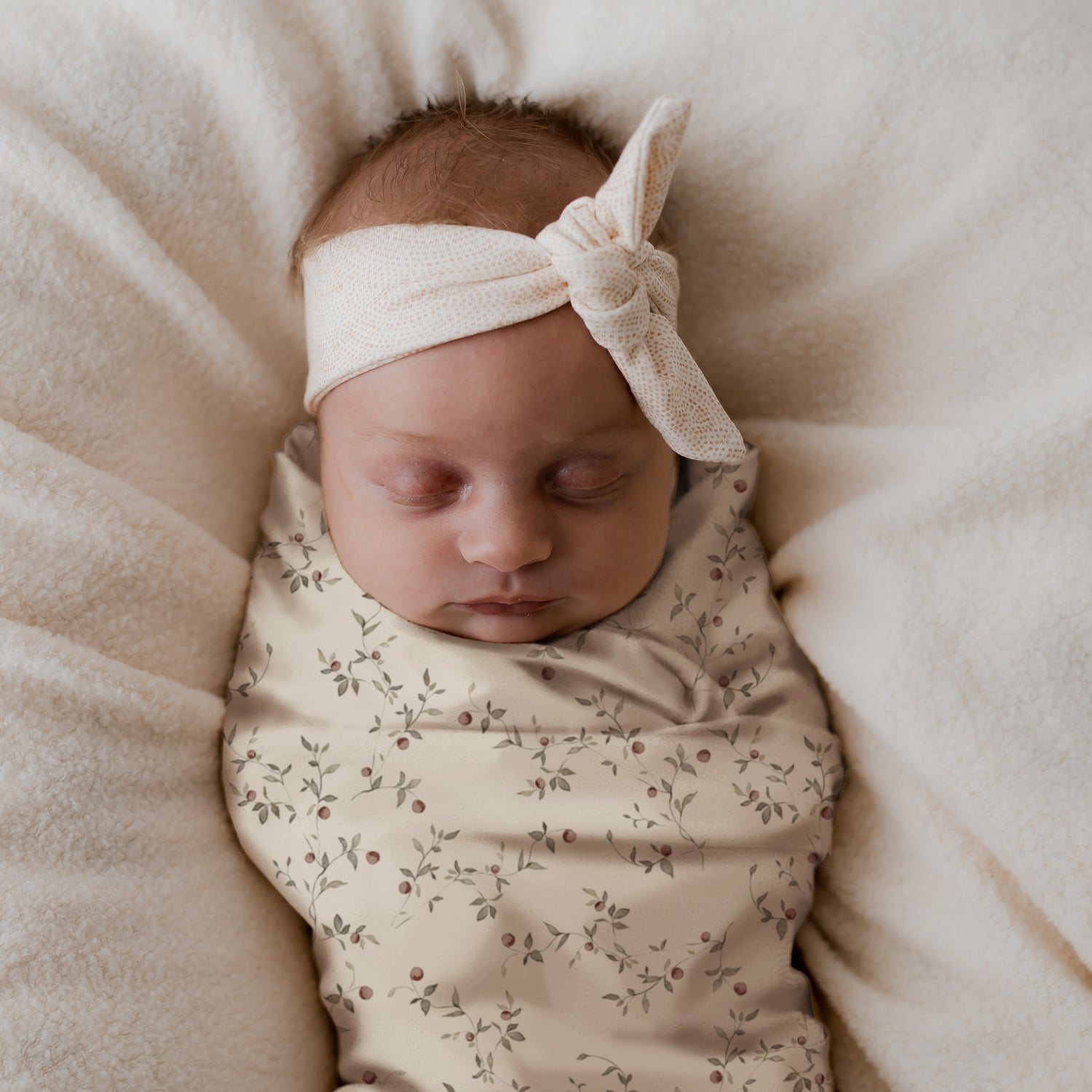 Een vredige pasgeboren baby ingebakerd in een Inbakerdoek Apple Branches wikkeldoek met strikhoofdband, rustend op een zachte, romige deken.