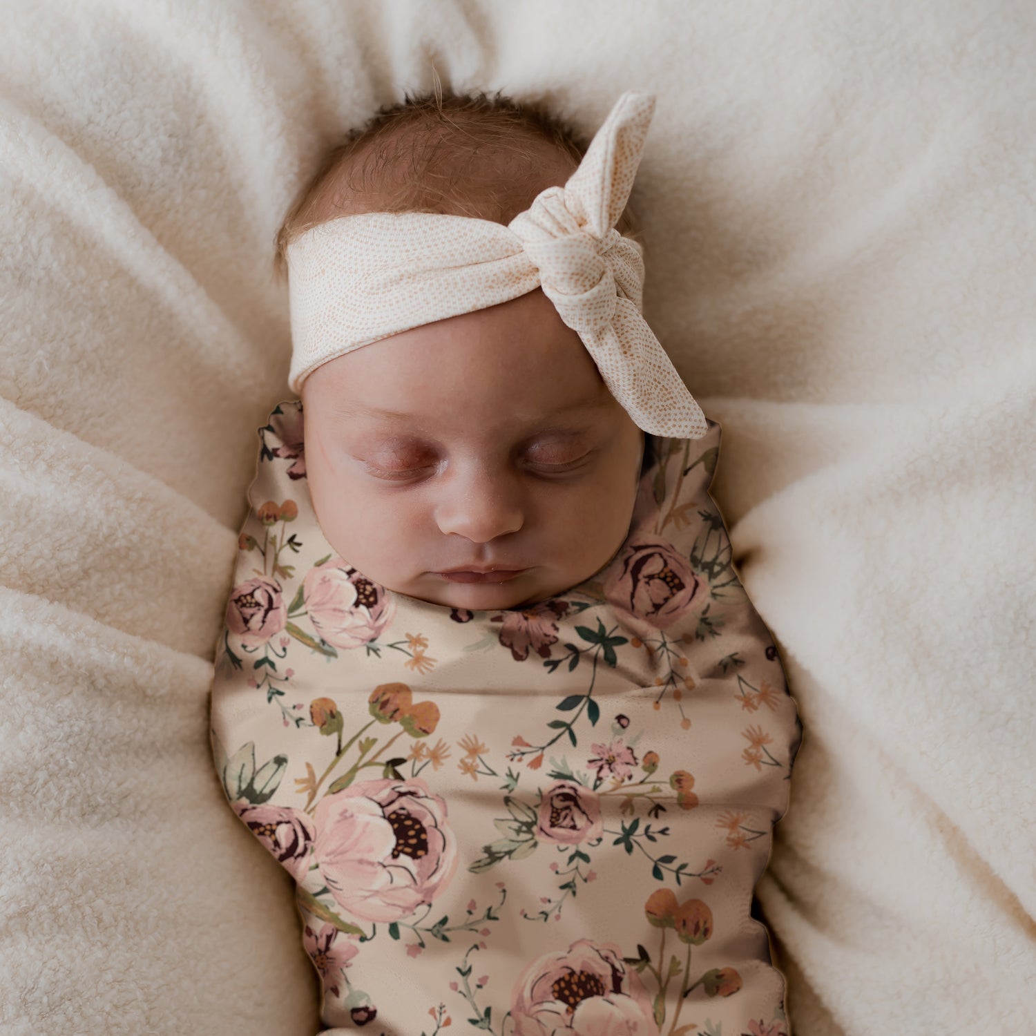 Een vredige pasgeborene ingebakerd in een gebloemde Inbakerdoek Blamy van Yumi Baby, met een strikhoofdband, sereen rustend op een zacht, crèmekleurig oppervlak.