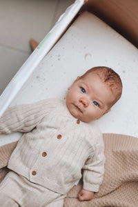 Een serene baby met betoverende blauwe ogen die comfortabel in een co-sleeper ligt, gekleed in een gezellig beige outfit gemaakt van Oeko-Tex katoen, lijkt de mysteries van het leven te overdenken met een Yumi Baby Wieghoeslaken Galaxy.