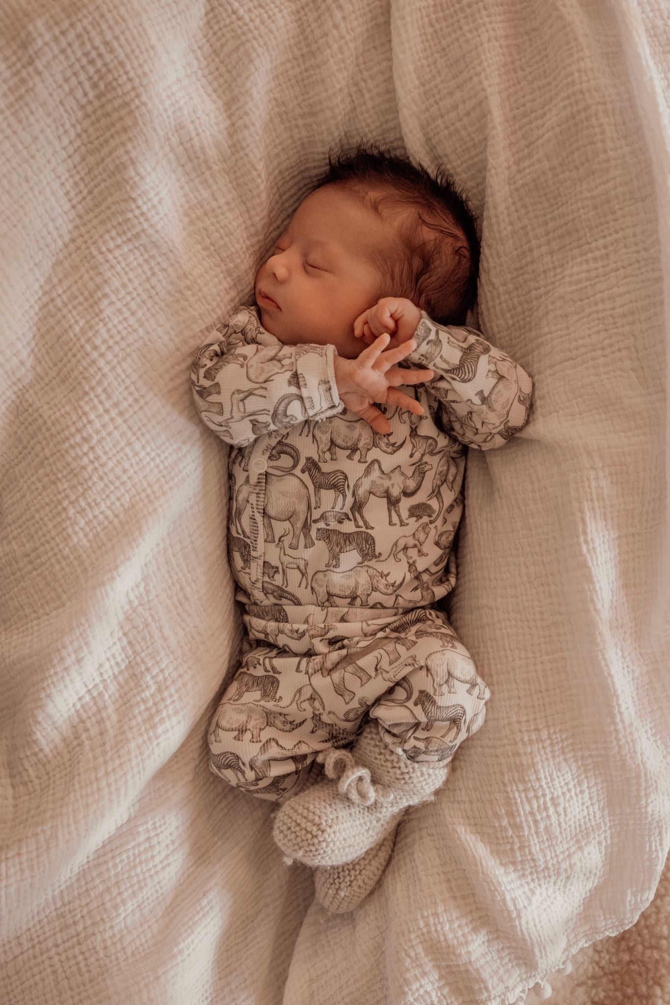 Een gezellige baby gekleed in een knus Yumi Baby Broekje Little Safari-outfit met een schattig bijpassend hoedje, ligt in een geweven mand, omringd door zachte, verkreukelde dekens, tevreden en vredig.