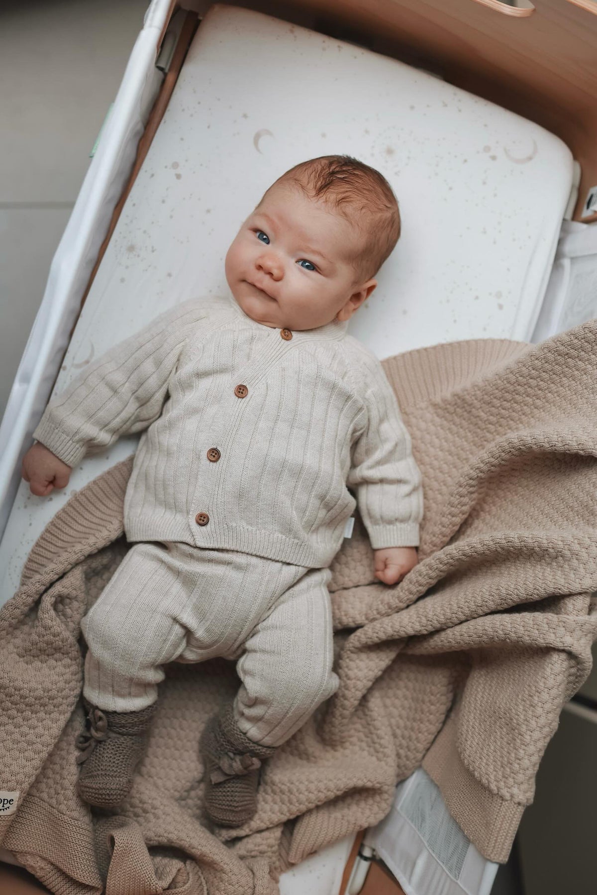Een serene baby met betoverende blauwe ogen die comfortabel in een co-sleeper ligt, gekleed in een gezellig beige outfit gemaakt van Oeko-Tex katoen, lijkt de mysteries van het leven te overdenken met een Yumi Baby Wieghoeslaken Galaxy.