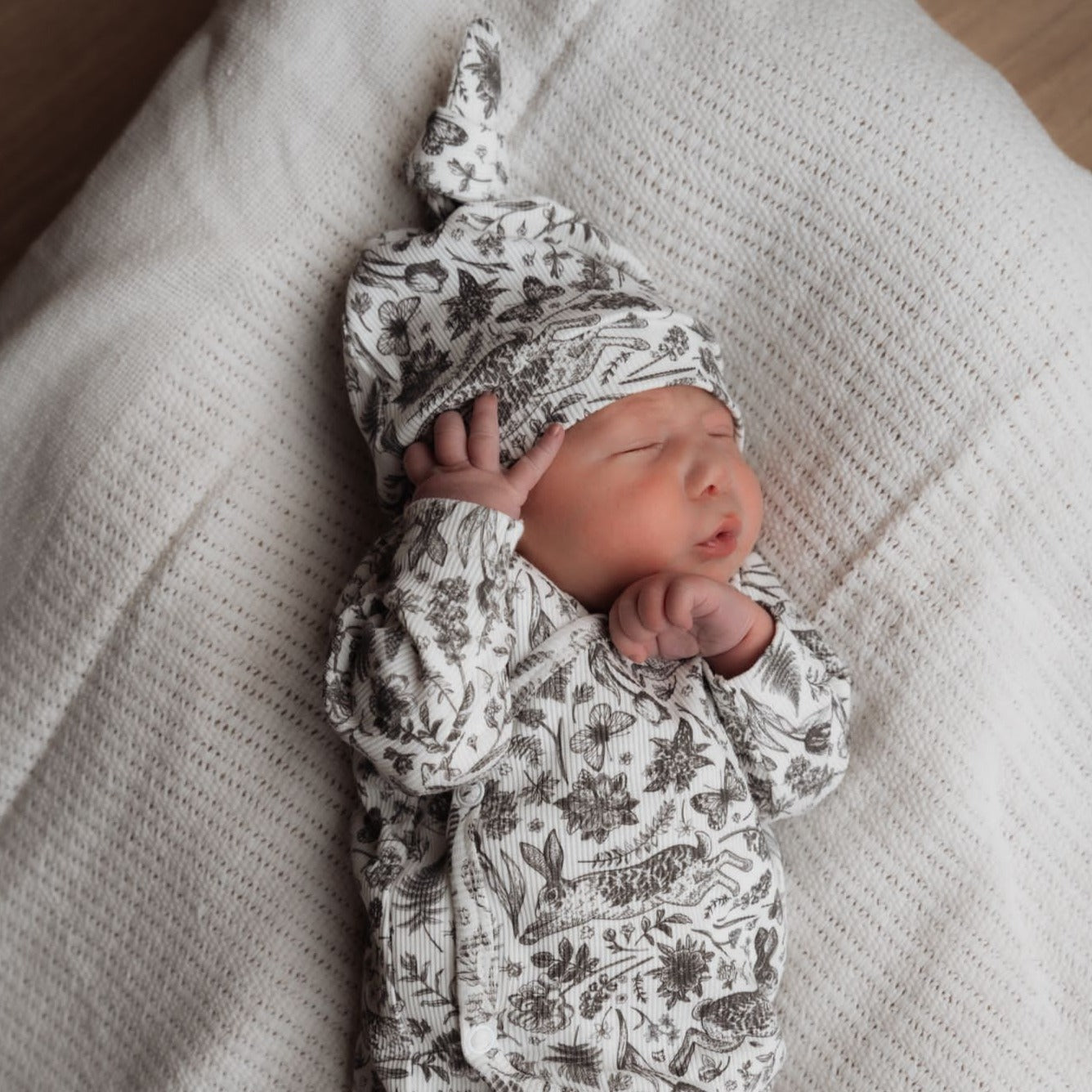 Een stille pasgeboren baby, gehuld in een babyoutfit met patronen en een Muts Bunnies van het merk Osman, slaapt zacht op een zachte, witte deken.