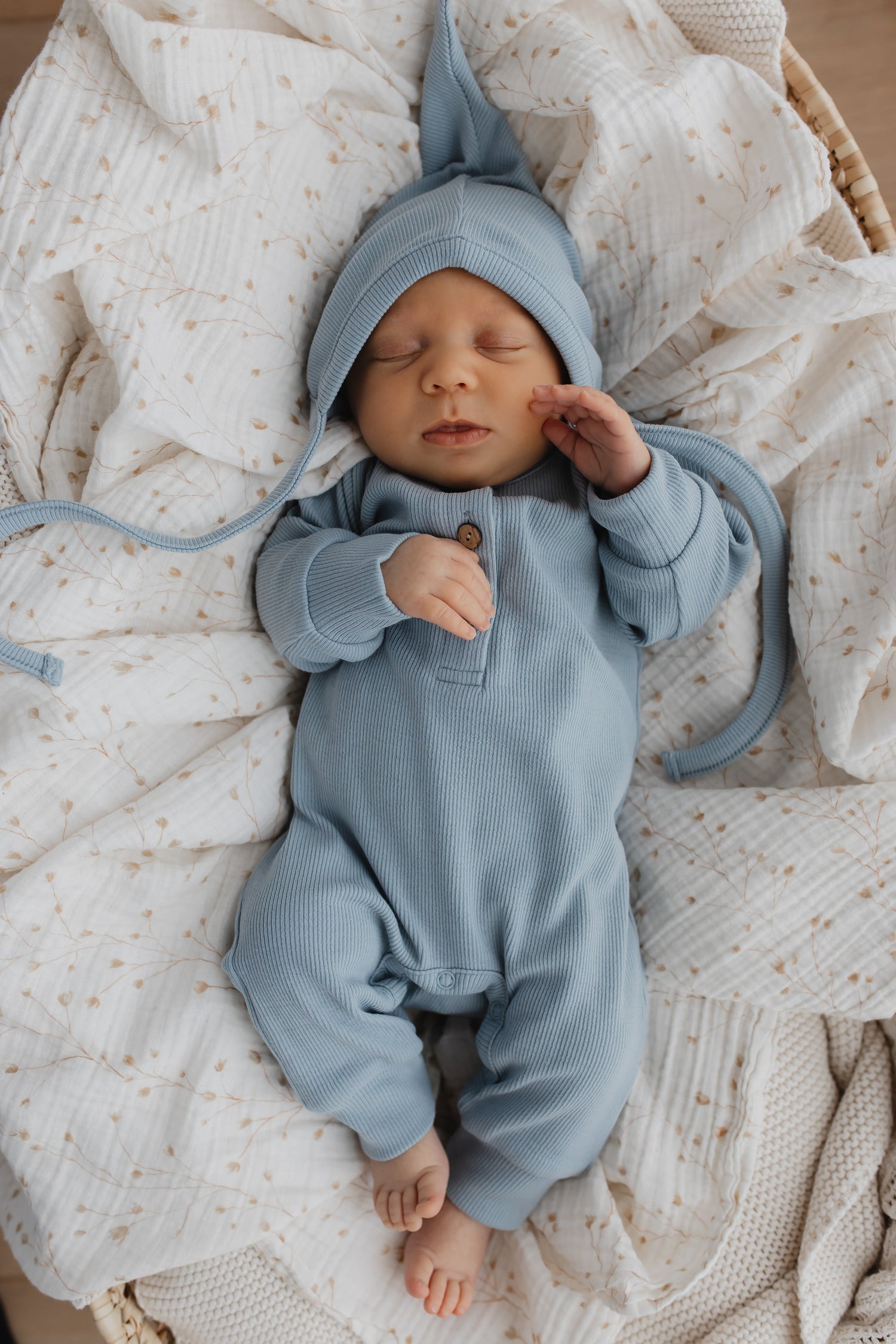 Een vredige pasgeboren baby slaapt heerlijk, gekleed in een Yumi Baby Boxpakje Blue Sky van hoogwaardig katoen met bijpassend mutsje, genesteld in een zacht crèmekleurig dekentje.