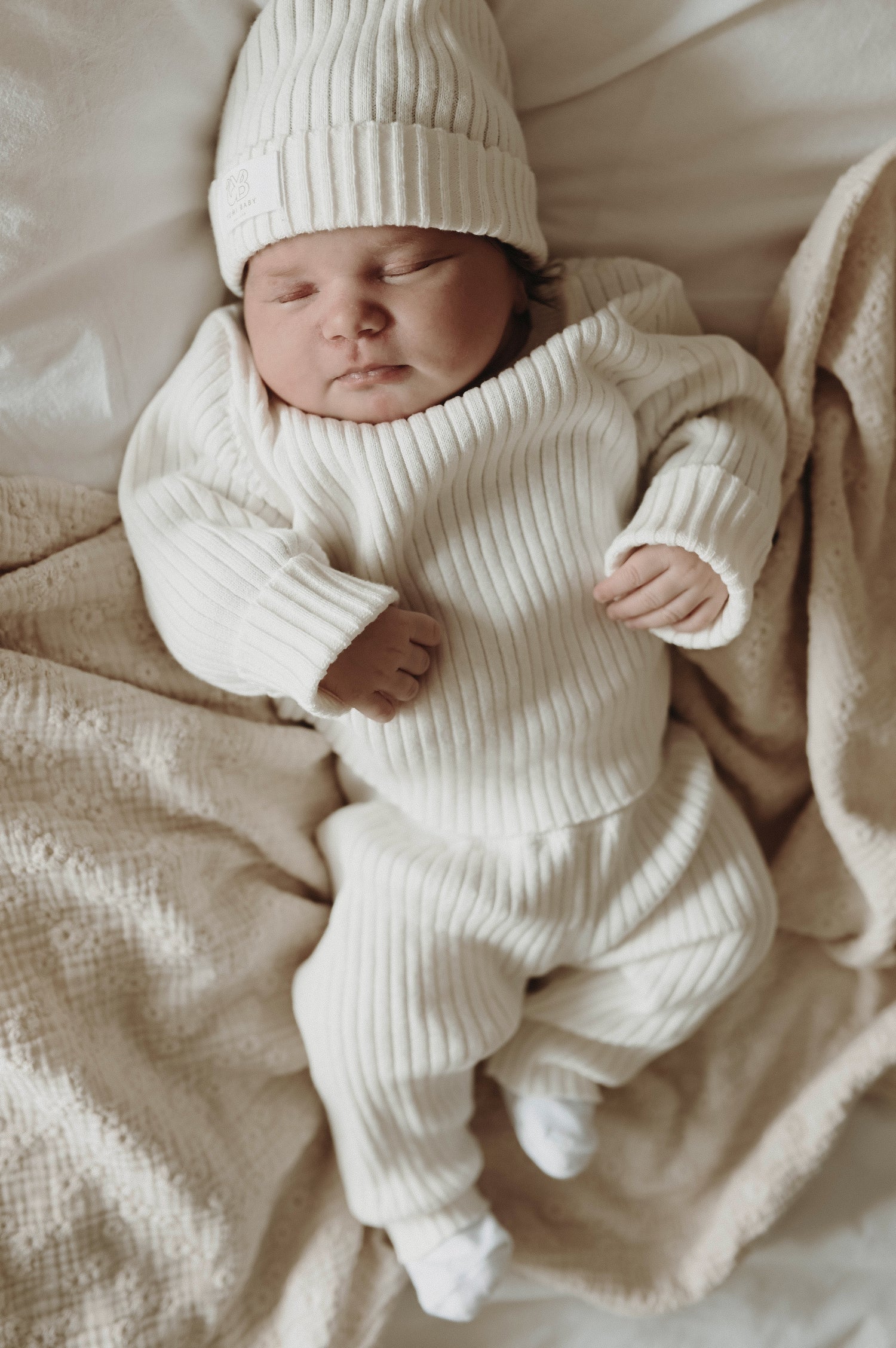 Een vredige pasgeboren baby, gekleed in witte kleding, lag comfortabel in een klein rotan wiegje, met een zachte deken en een delicate Yumi Baby Knitted Muts Pearl Whisper 0-3 mnd zachtjes gedrapeerd over de zijkant.