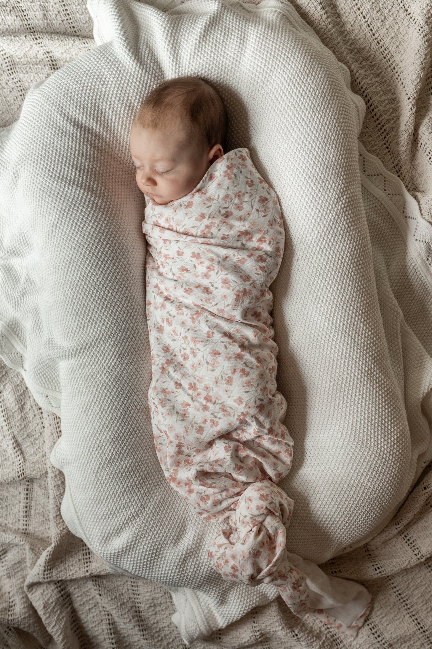 Een vredige pasgeboren baby, knus gewikkeld in een Yumi Baby Inbakerdoek Fleur de Jardin, heerlijk slapend op een zachte, gestructureerde deken.