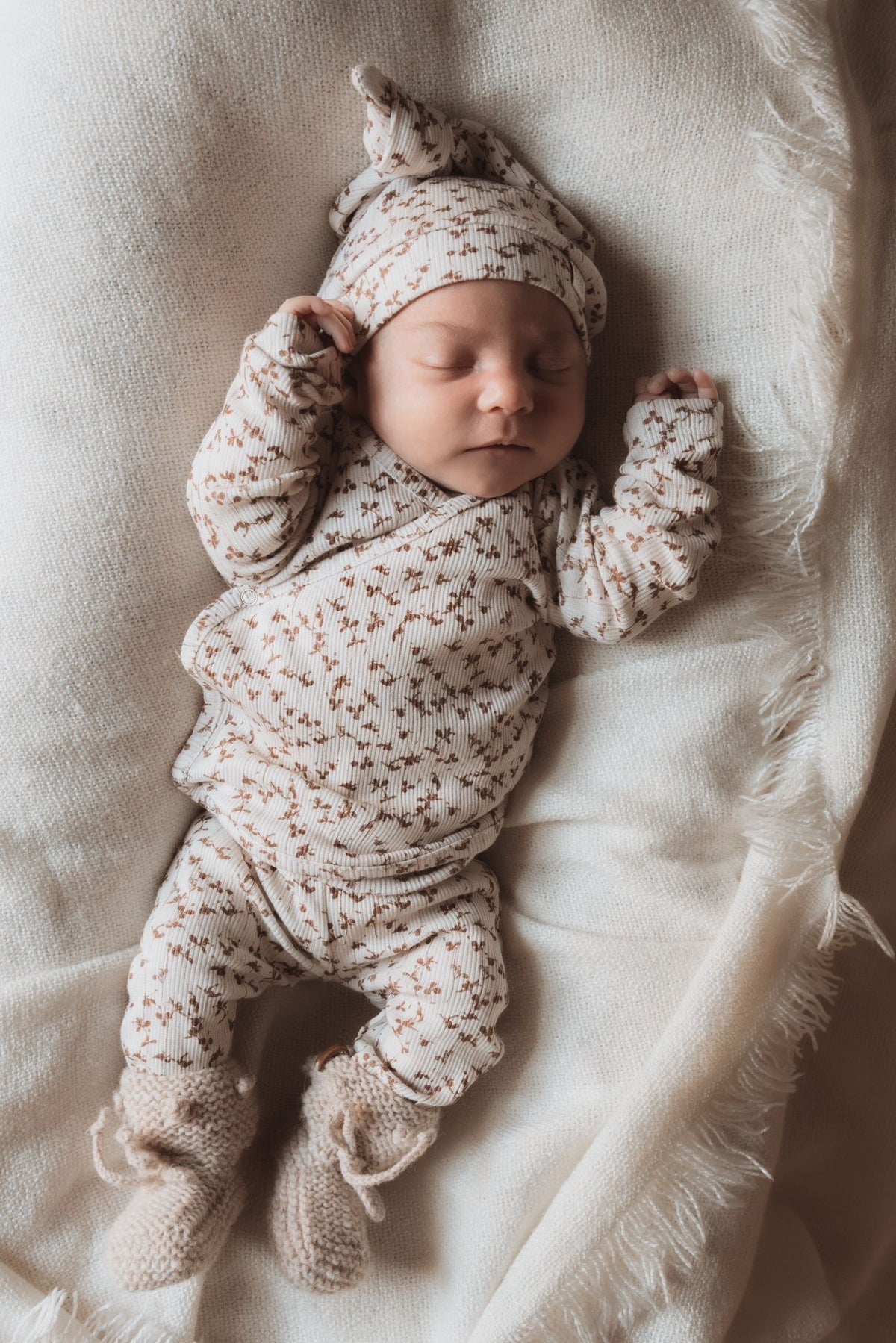 Een vredige pasgeborene slaapt heerlijk, gekleed in een Yumi Baby Overslagshirt Brown Leafs met bijpassend mutsje en slofjes, allemaal gemaakt van OEKO TEX katoen, genesteld op een zachte, crèmekleurige deken.