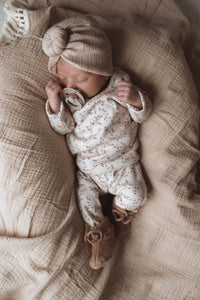 Een vredige pasgeboren baby gekleed in een Yumi Baby Overslagshirt Apple Branches, gemaakt van OEKO TEX-katoen, met bijpassend mutsje en kleine slofjes, slaapt heerlijk op een zachte, gestructureerde deken en clutch.