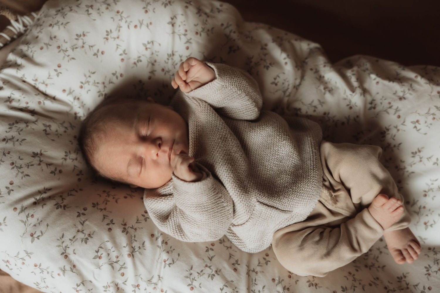 Een pasgeboren baby slaapt vredig in een Yumi Baby co-sleeper, gekleed in een beige trui en lichtbruine broek, rustend op een zacht patroon Wieghoeslaken Appeltakken kussen.