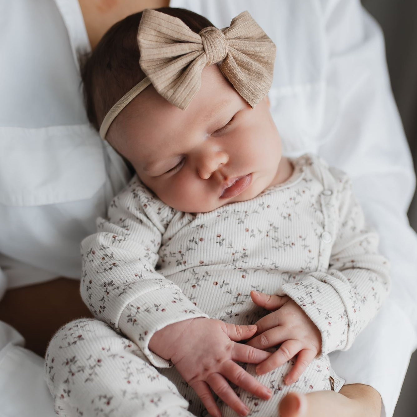 Een vredige pasgeborene slaapt heerlijk in een Yumi Baby Boxpakje Appeltakken, gewiegd in een liefdevolle omhelzing, versierd met een schattige strikjeshoofdband.