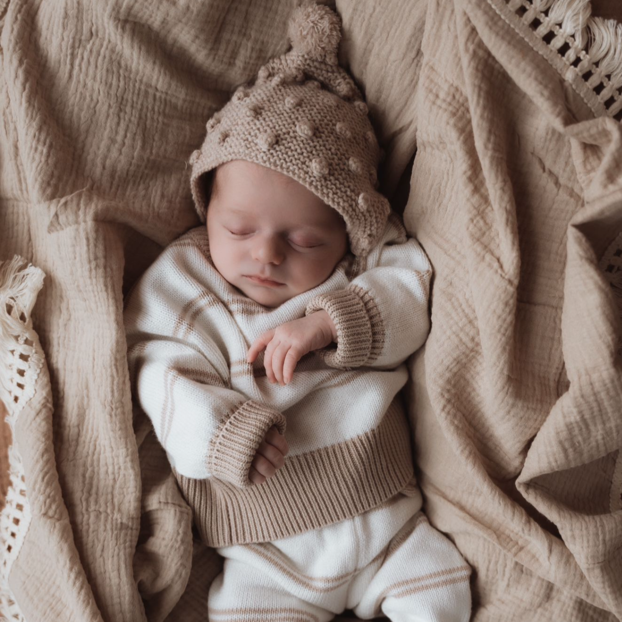 Een vredige pasgeboren baby gaapt terwijl hij comfortabel in een geweven wiegje ligt, gewikkeld in een zachte 100% Yumi Baby gebreide broek Sandy Stripe deken.