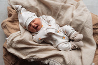 Een vredige pasgeborene, gekleed in een Yumi Baby Boxpakje Oranjebeige onesie met een bijpassend mutsje, rustend in een knus geweven mandje, gevoerd met een zachte witte deken.