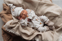 Een vredige pasgeborene, gekleed in een Yumi Baby Boxpakje Oranjebeige onesie met een bijpassend mutsje, rustend in een knus geweven mandje, gevoerd met een zachte witte deken.