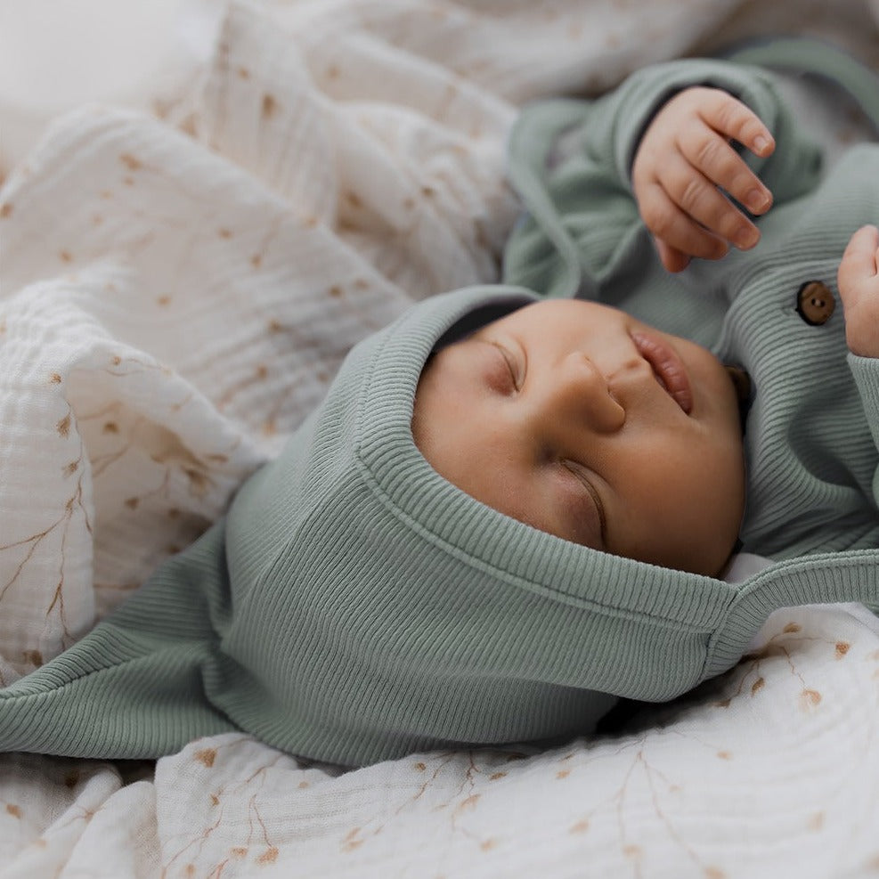 Een pasgeboren baby slaapt emotioneel, ingewikkeld in een zacht Strikmutsje Jade Treasure van Yumi Baby, gemaakt van bestendige materialen en rustend op gedragsmatige dekens.