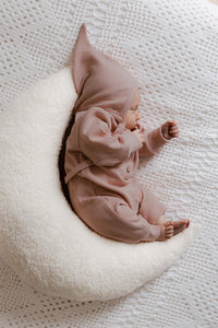Een vredig kindje gekleed in een gezellig bruin Boxpakje Blush gemaakt van hoogwaardig katoen van Yumi Baby, met een bijpassend hoedje, opgerold en duttend op een zacht wit halvemaanvormig kussen.