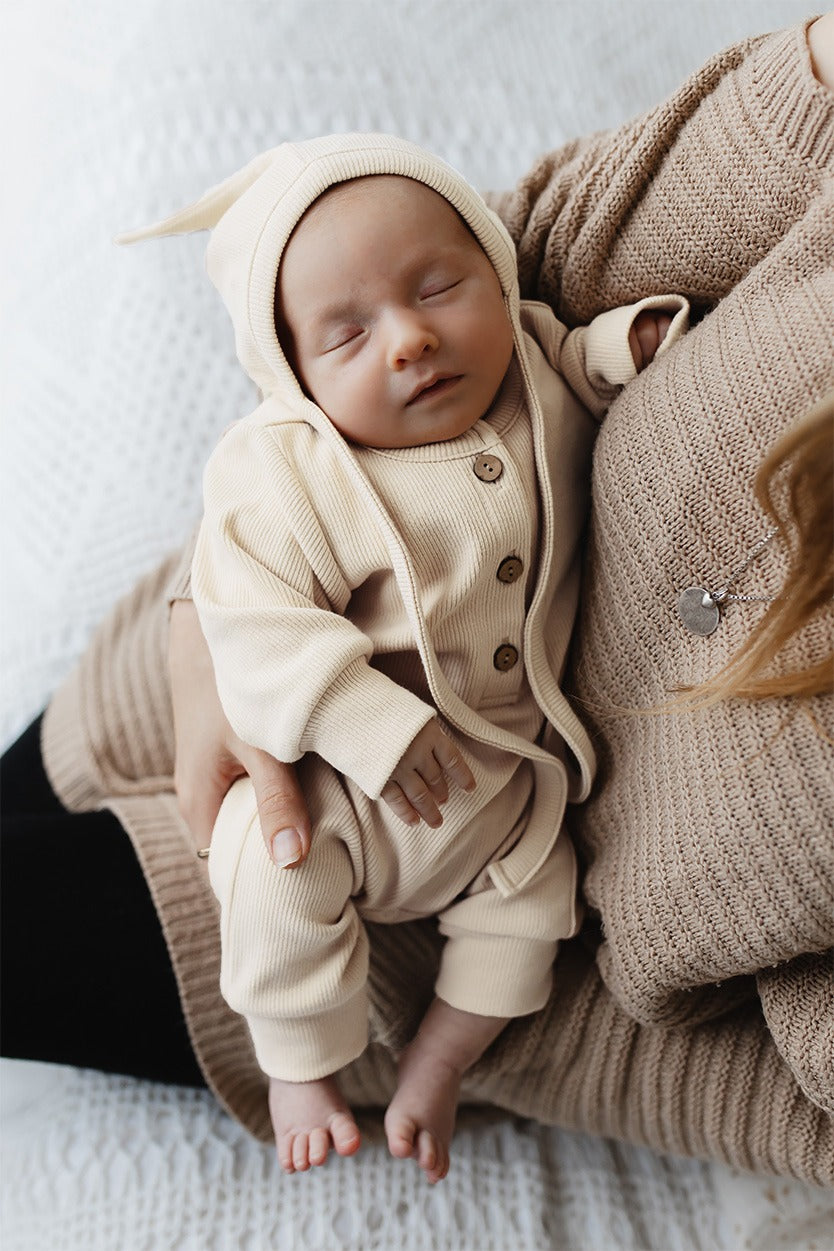 Vreedzaam slapen: een pasgeboren baby slaapt heerlijk, gewiegd door de zachte aanraking van een voogd, gekleed in een gezellige crèmekleurige outfit met schattige capuchon en Strikmutsje Ivory Dreams van Yumi Baby.