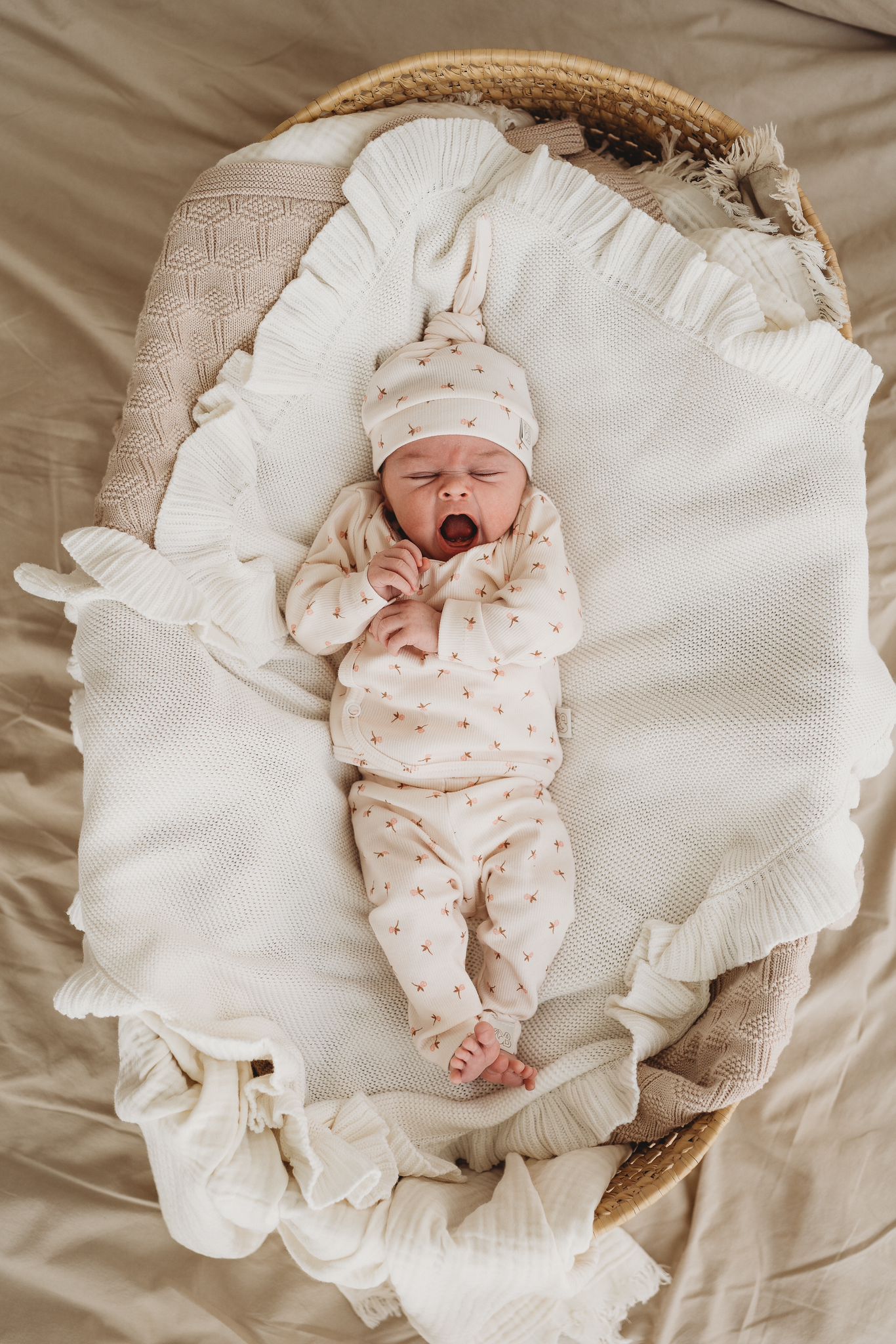Een gapende baby gekleed in een knus rompertje met een bijpassend pasgeboren Muts Tiny Tulip babymutsje van Yumi Baby, comfortabel genesteld in een geweven mand op een zachte, zijdezachte deken.