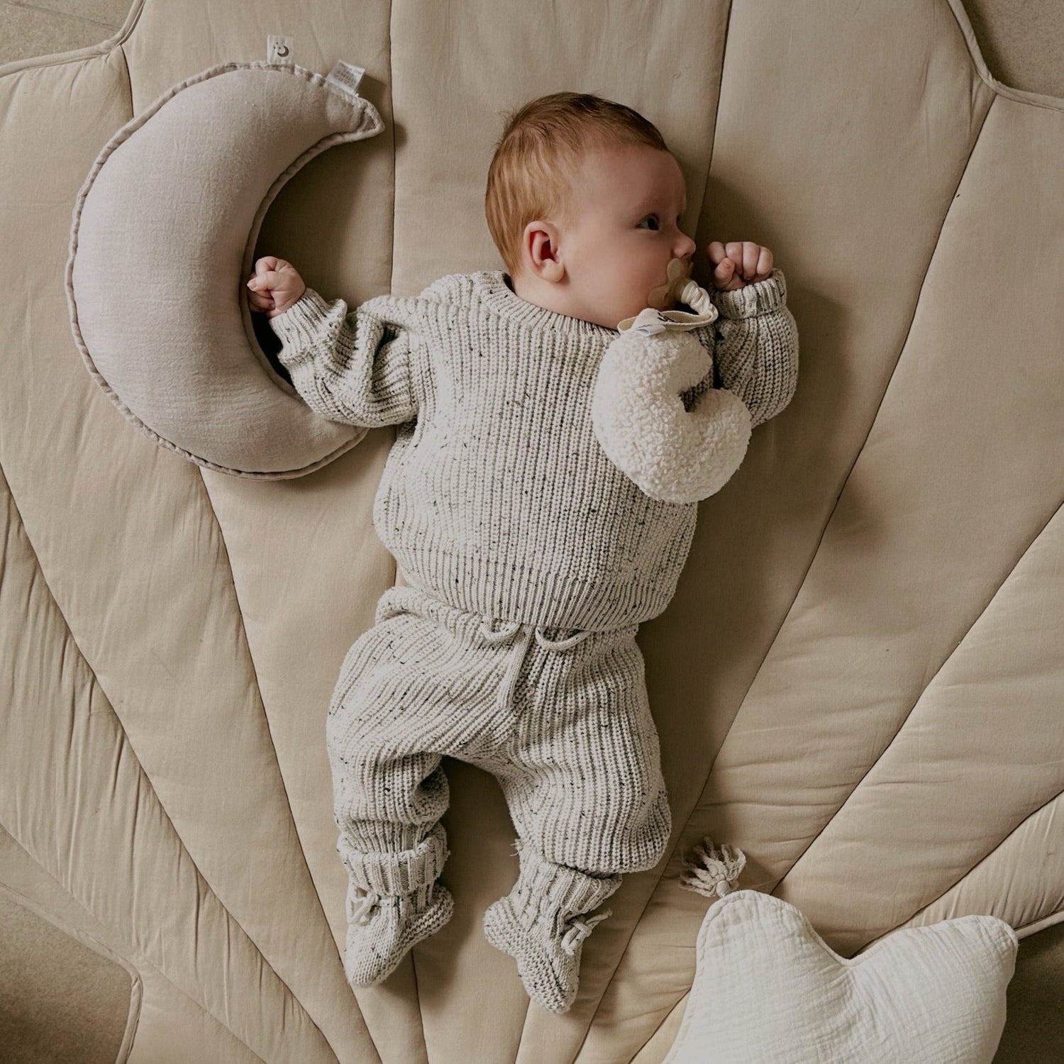 Een vredige baby gekleed in een gezellige Yumi Baby Knitted Set Salt & Pepper-outfit, liggend op een zachte, neutraal gekleurde deken, met een halvemaanvormig kussen in de buurt.