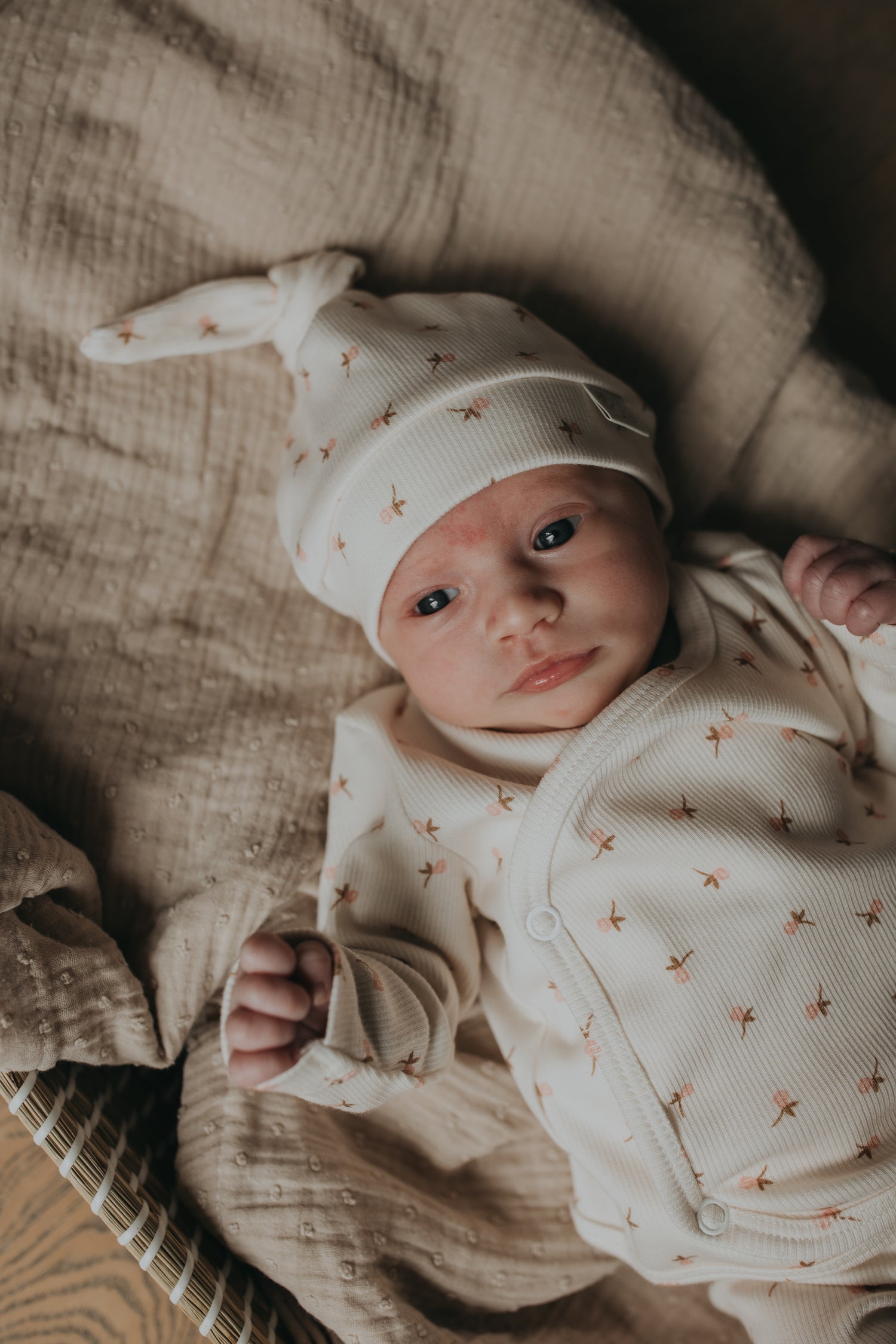 Een gapende baby gekleed in een knus rompertje met een bijpassend pasgeboren Muts Tiny Tulip babymutsje van Yumi Baby, comfortabel genesteld in een geweven mand op een zachte, zijdezachte deken.