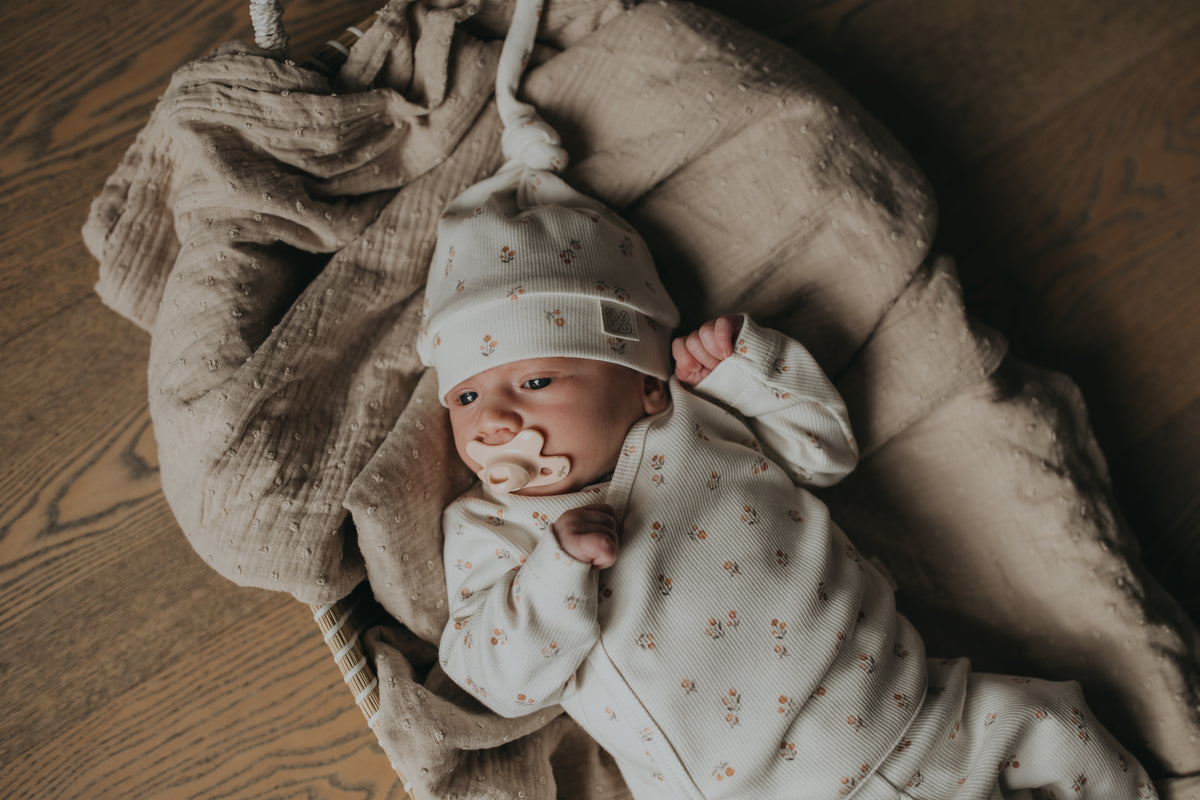 Een vredige pasgeboren baby in een schattig wit outfitje met printjes, wegdommelend met kleine handjes bij het gezichtje, lekker weggestopt op een zacht, zijdezacht dekentje met een Yumi Baby Muts Peach Blossom op het hoofdje.