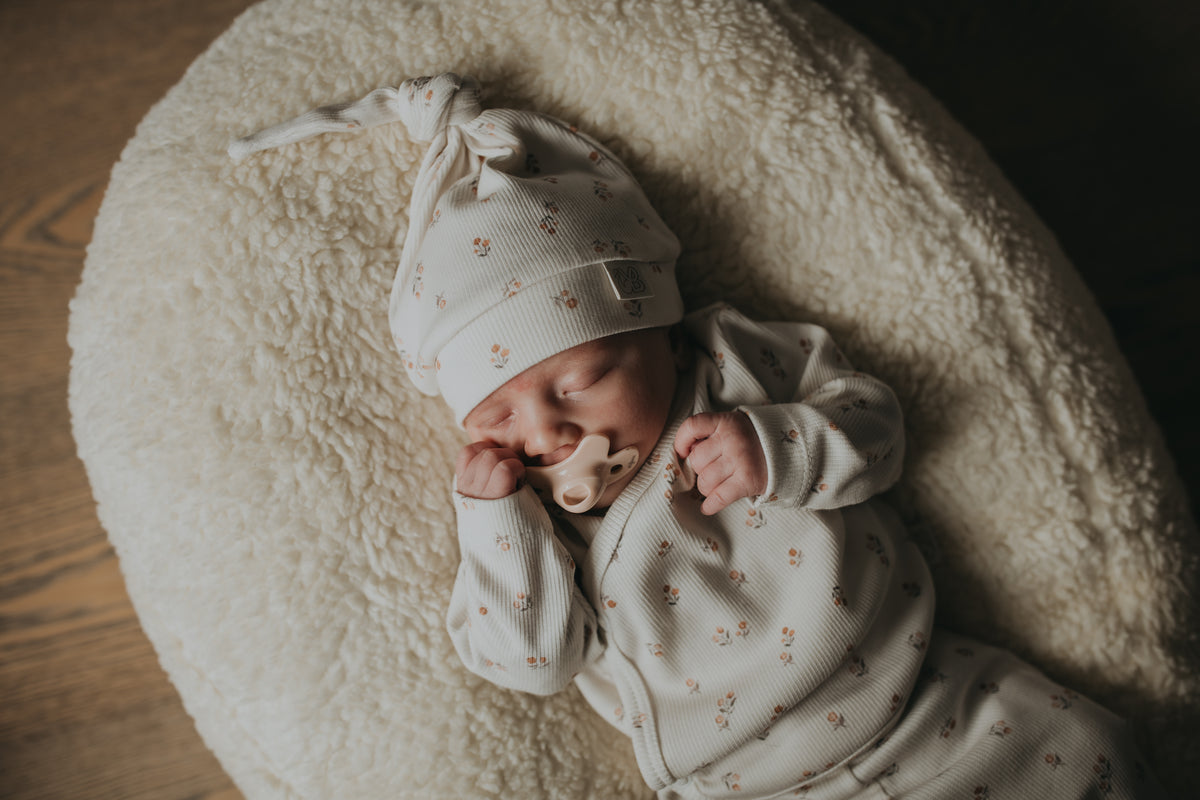 Een vredige pasgeboren baby in een schattig wit outfitje met printjes, wegdommelend met kleine handjes bij het gezichtje, lekker weggestopt op een zacht, zijdezacht dekentje met een Yumi Baby Muts Peach Blossom op het hoofdje.