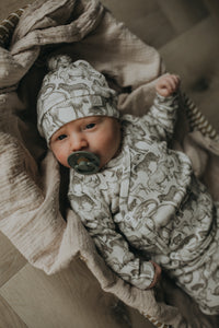 Een gezellig uitziende baby, gekleed in een Yumi Baby Overslagshirt Little Safari-onesie en bijpassend mutsje, rustend in een geweven mand op een zachte, milieuvriendelijke deken.