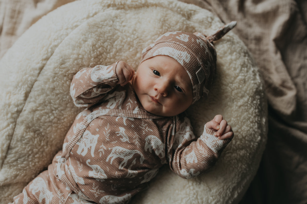 Een vredige pasgeboren baby gekleed in een rompertje met patroon en bijpassende Muts Boho Jungle, heerlijk slapend in een knus mandje omringd door zachte, crèmekleurige dekens.
