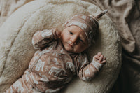 Een vredige pasgeboren baby gekleed in een rompertje met patroon en bijpassende Muts Boho Jungle, heerlijk slapend in een knus mandje omringd door zachte, crèmekleurige dekens.