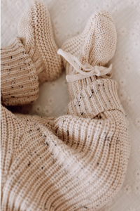 Een paar knusse, 100% katoenen Yumi Baby handgebreide beige slofjes, waarschijnlijk voor een baby van 0-12 maanden, vastgebonden met delicate strikjes, die zachtjes tegen elkaar rusten.