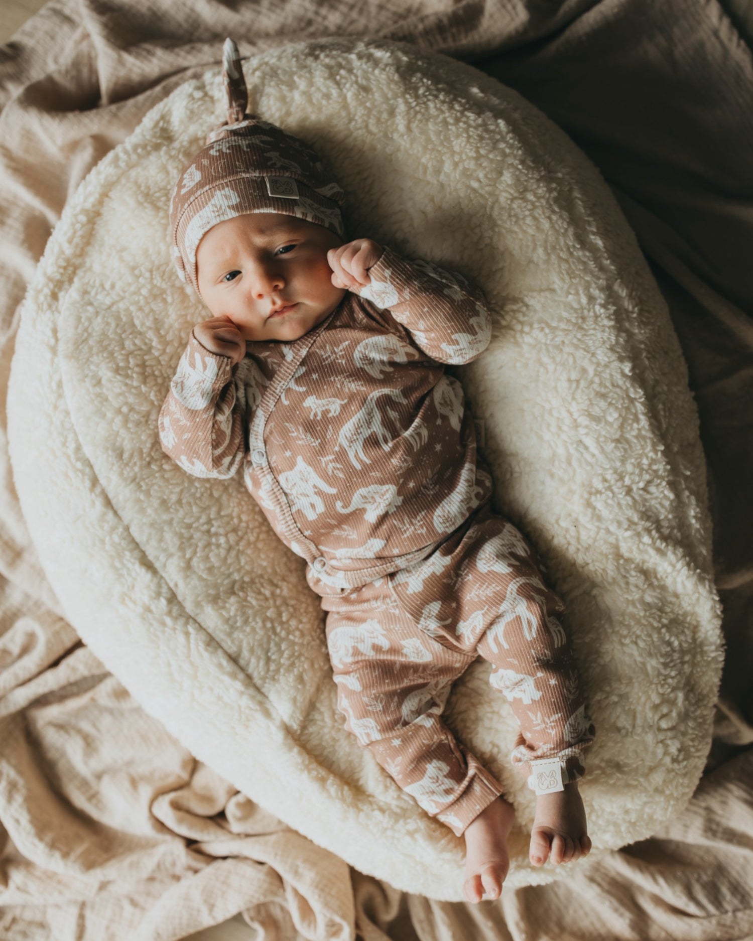 Een vredige pasgeboren baby gekleed in een rompertje met patroon en bijpassende Muts Boho Jungle, heerlijk slapend in een knus mandje omringd door zachte, crèmekleurige dekens.