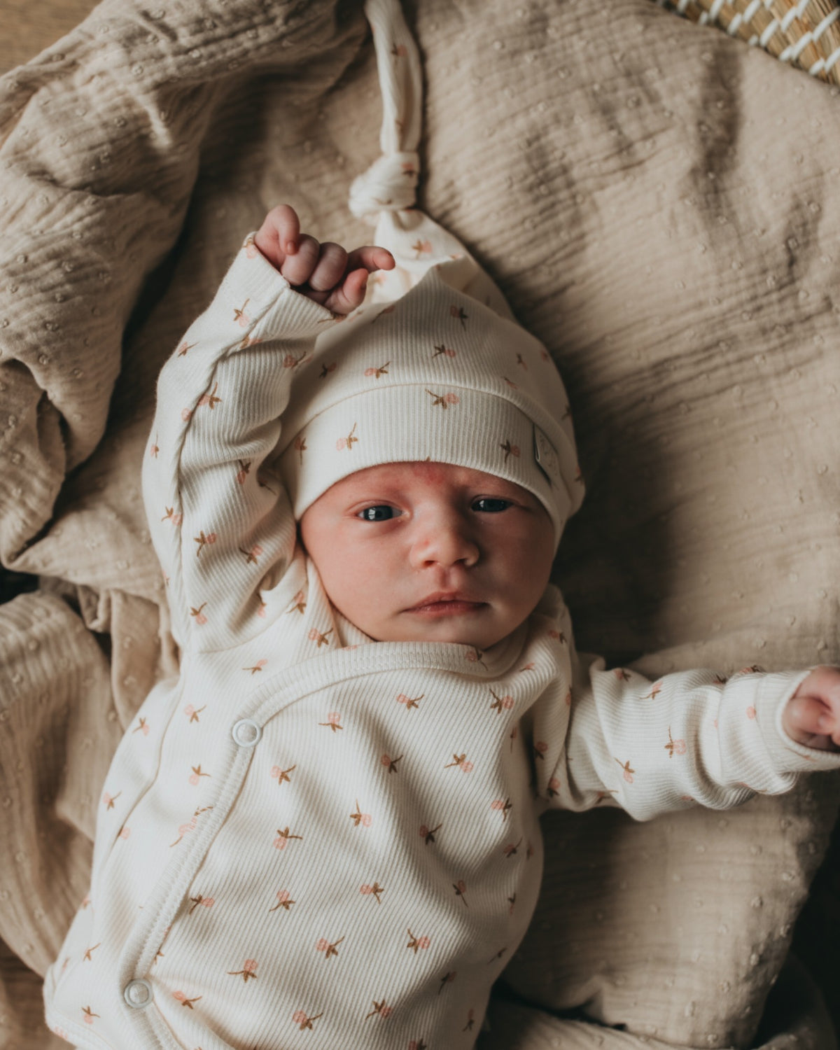 Een gapende baby gekleed in een knus rompertje met een bijpassend pasgeboren Muts Tiny Tulip babymutsje van Yumi Baby, comfortabel genesteld in een geweven mand op een zachte, zijdezachte deken.