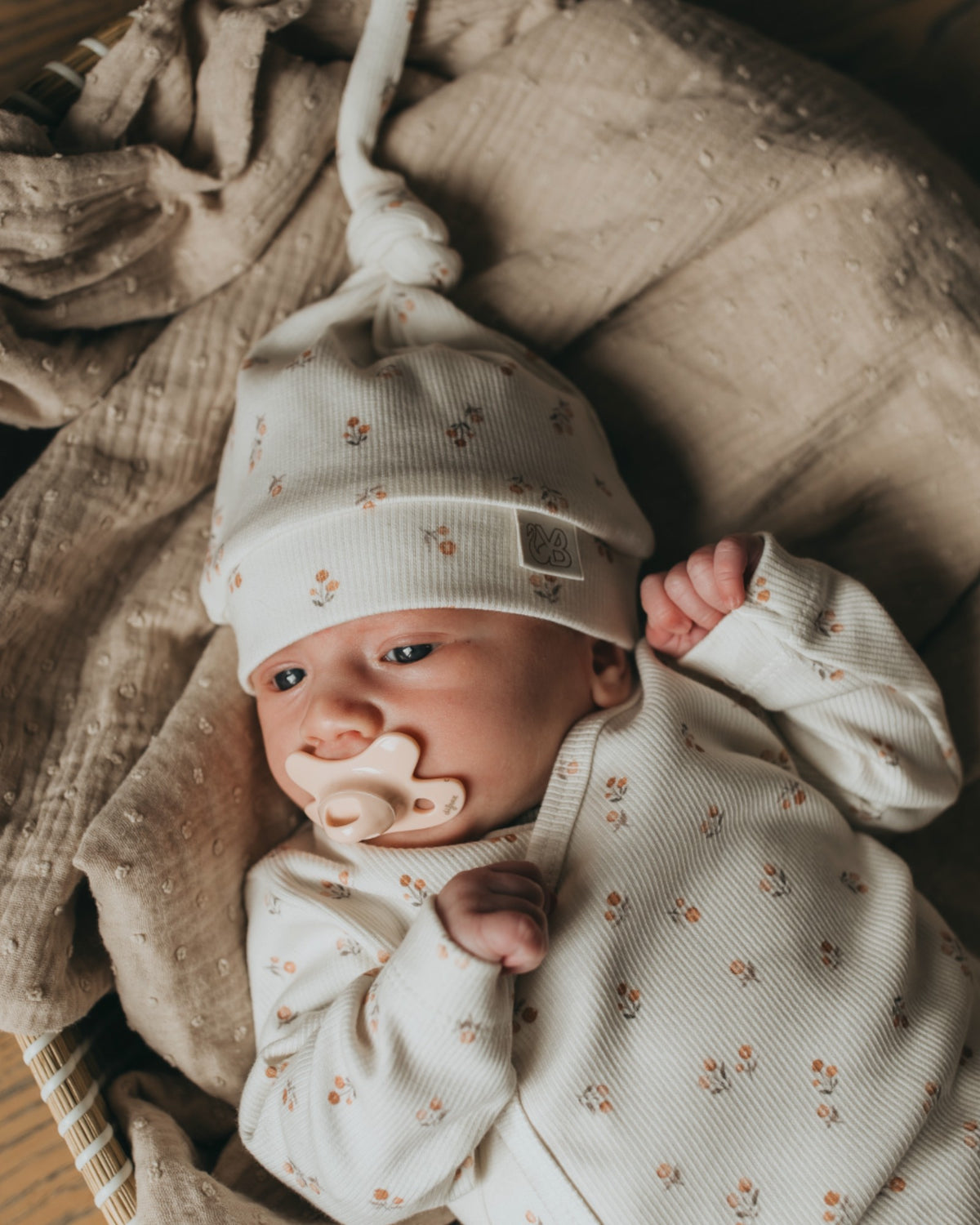 Een vredige pasgeboren baby in een schattig wit outfitje met printjes, wegdommelend met kleine handjes bij het gezichtje, lekker weggestopt op een zacht, zijdezacht dekentje met een Yumi Baby Muts Peach Blossom op het hoofdje.