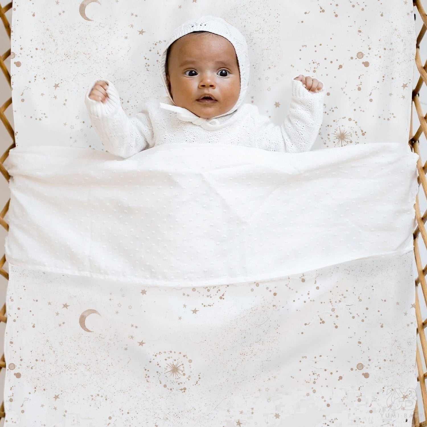 Een serene baby, weggestopt in een gezellig Wieglaken Galaxy versierd met hemelse patronen, genesteld in een geweven "wiegje" gemaakt van Oeko Tex-katoen, kijkt met grote ogen verbaasd op.