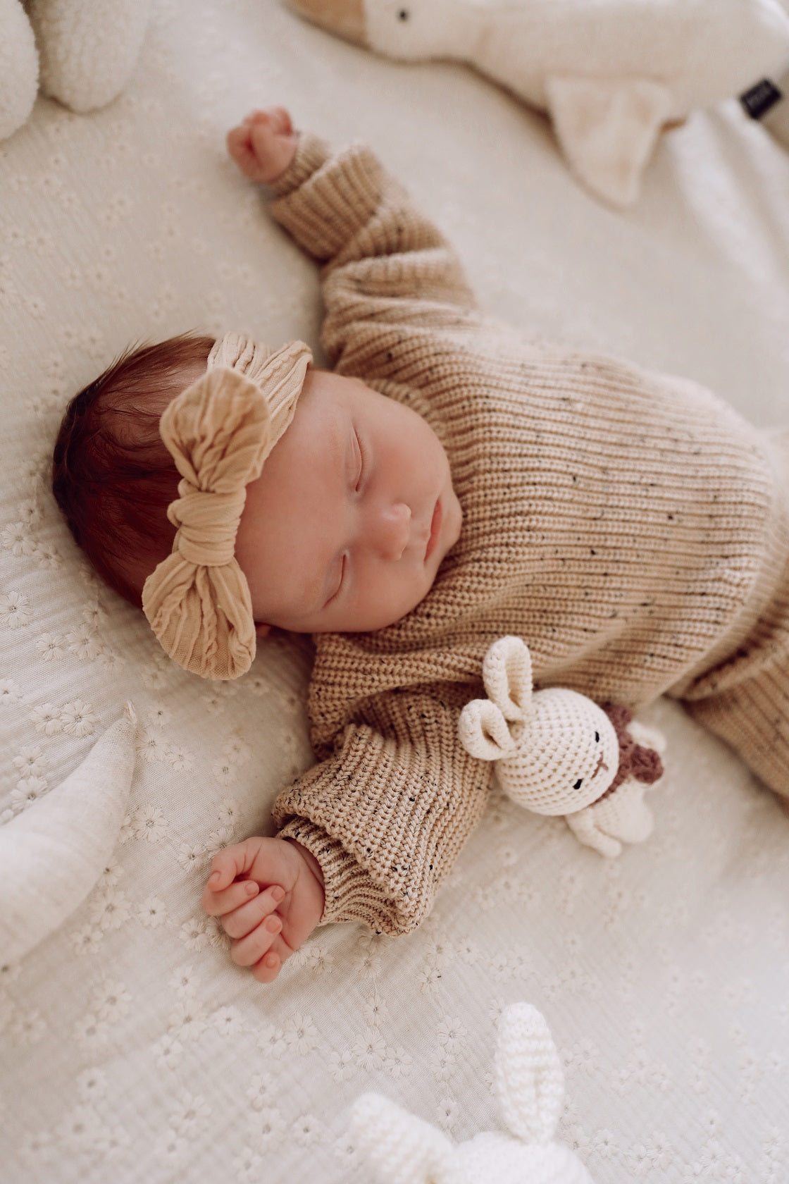 Een vredige baby slaapt heerlijk in een knus beige Yumi Baby Gebreide Set-outfit, met een zachte hoofdband en een klein knuffelkonijntje aan haar zijde.