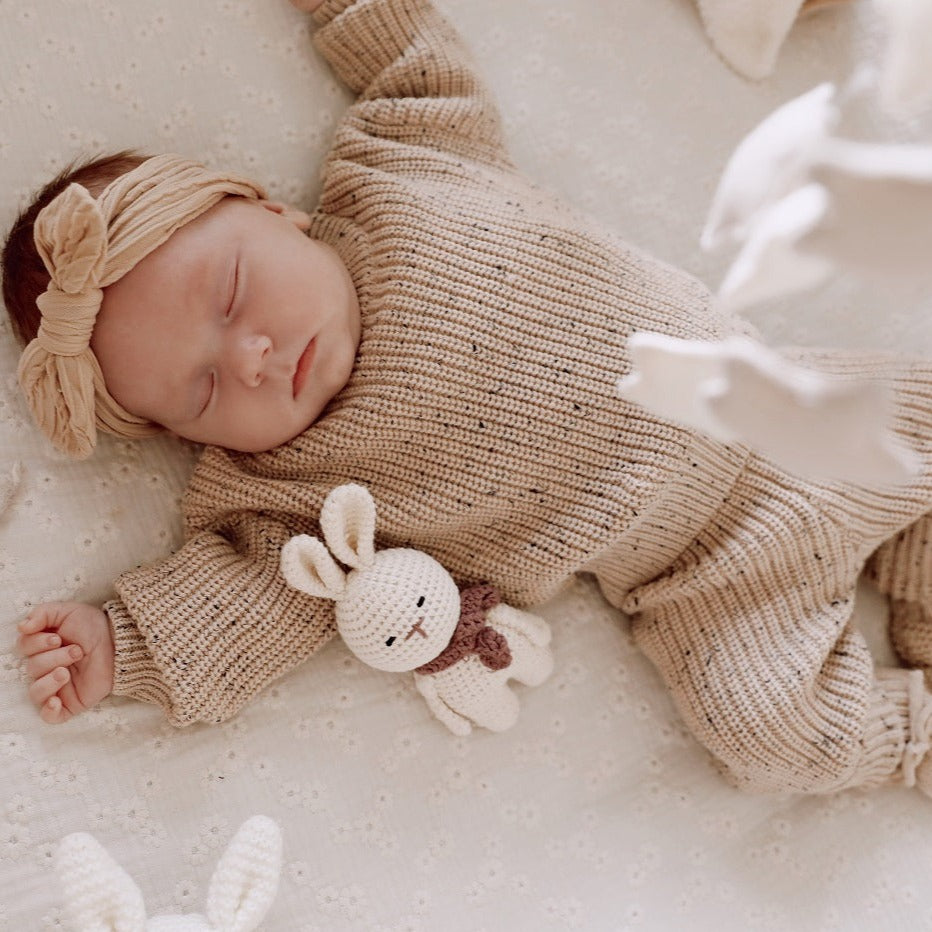 Een vredig slapende baby in een knus Yumi Baby 100% katoen Knitted Set Caramel outfit, met een zacht konijntje aan haar zijde, comfortabel genesteld op een zachte, neutraal getinte deken.