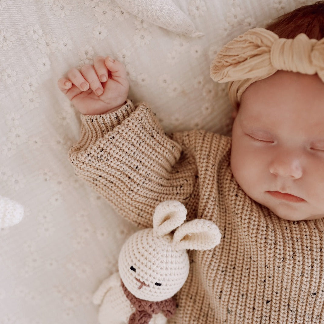 Een vredige baby slaapt heerlijk in een knus beige Yumi Baby Gebreide Set-outfit, met een zachte hoofdband en een klein knuffelkonijntje aan haar zijde.