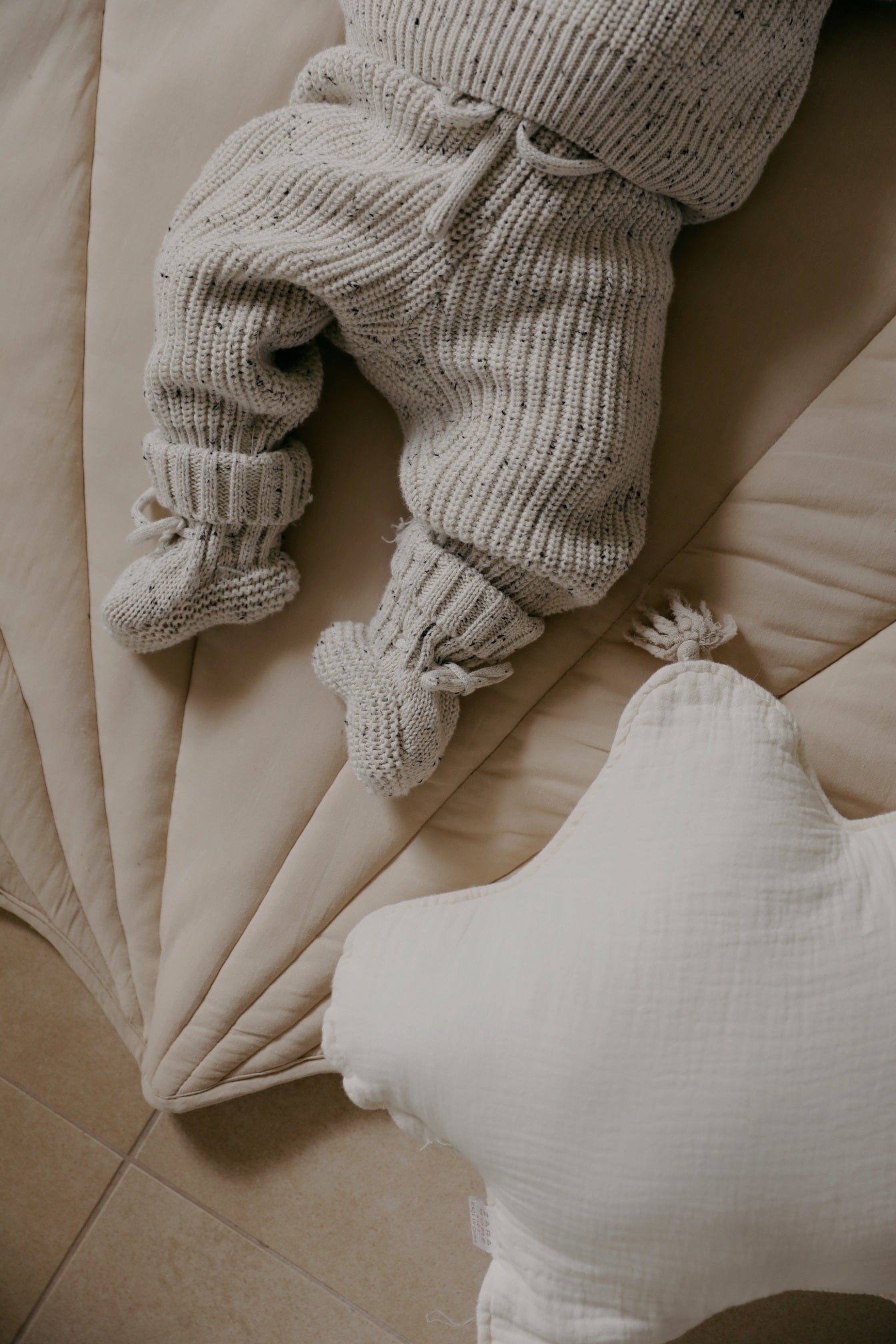 Gezellig en comfortabel, een kleintje gekleed in een Yumi Baby Knitted broek Salt & Pepper-outfit doet een vredig dutje op een zachte ondergrond, met een pluchen sterrenkussen en redelijke slofjes in de buurt.