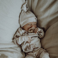 Een serene baby, gekleed in een Osman Broekje Vintage Jungle Taupe onesie en een knusse muts, rustig dommelend met een speentje, gehuld in een zachte, neutraal getinte deken gemaakt van duurzaam.