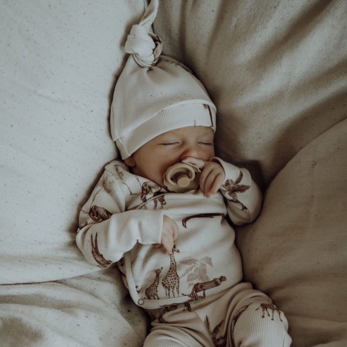 Een serene baby in een gezellig Yumi Baby Overslagshirt Vintage Jungle Taupe, compleet met bijpassend mutsje, rustig duttend met een speentje.
