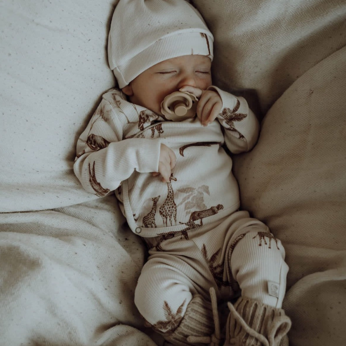 Een vredige pasgeboren baby, gekleed in een schattig outfitje met giraffenmotief en een mutsje, met een speentje, slaapt rustig op een zachte, OEKO-TEX gecertificeerde Yumi Baby Broekje Vintage Jungle Taupe deken.