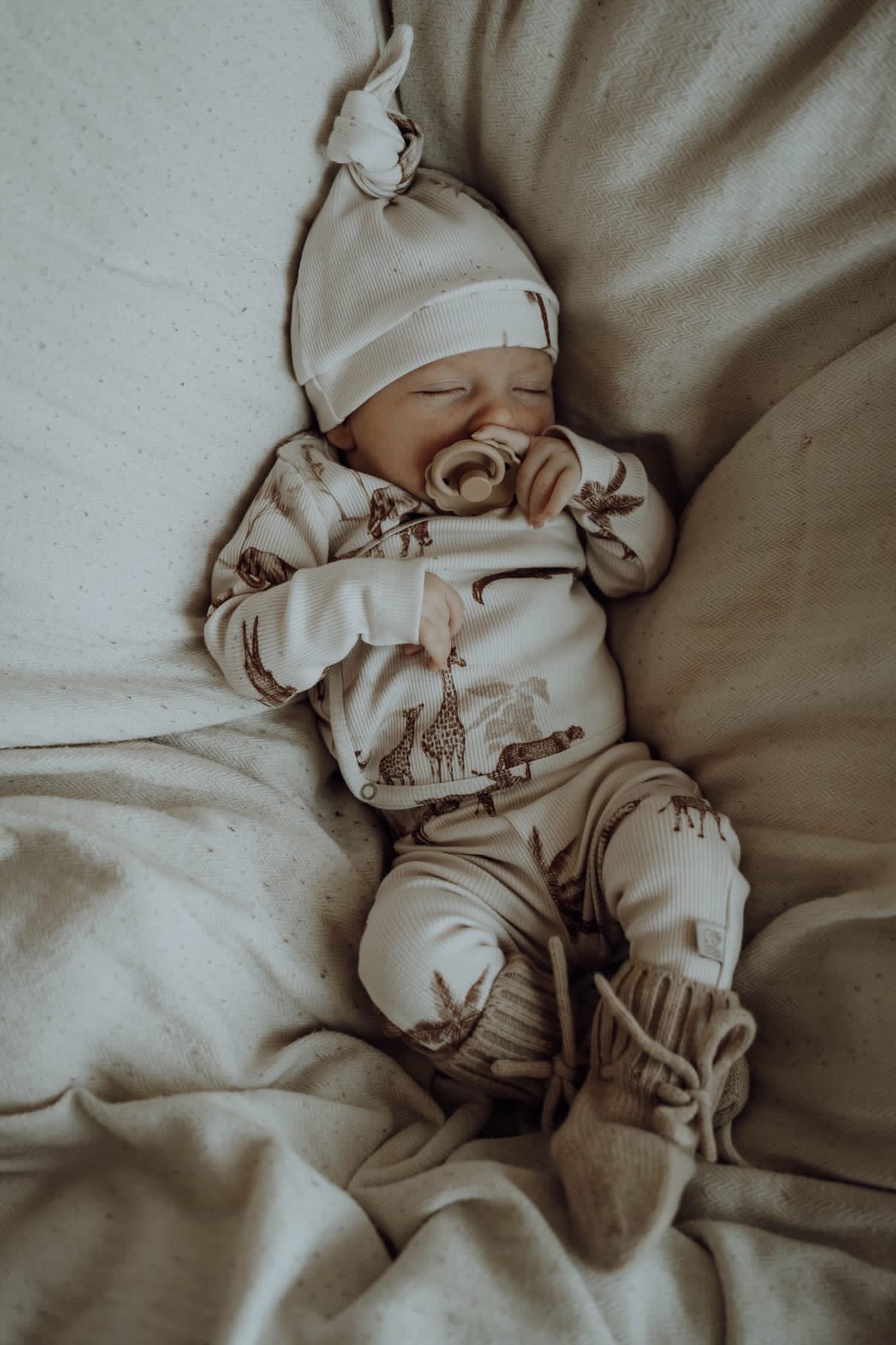 Een vredige newborn met een speentje die wegdommelt in een knus Osman Overslagshirt Vintage Jungle Taupe outfit, versierd met een schattig geknoopt mutsje.