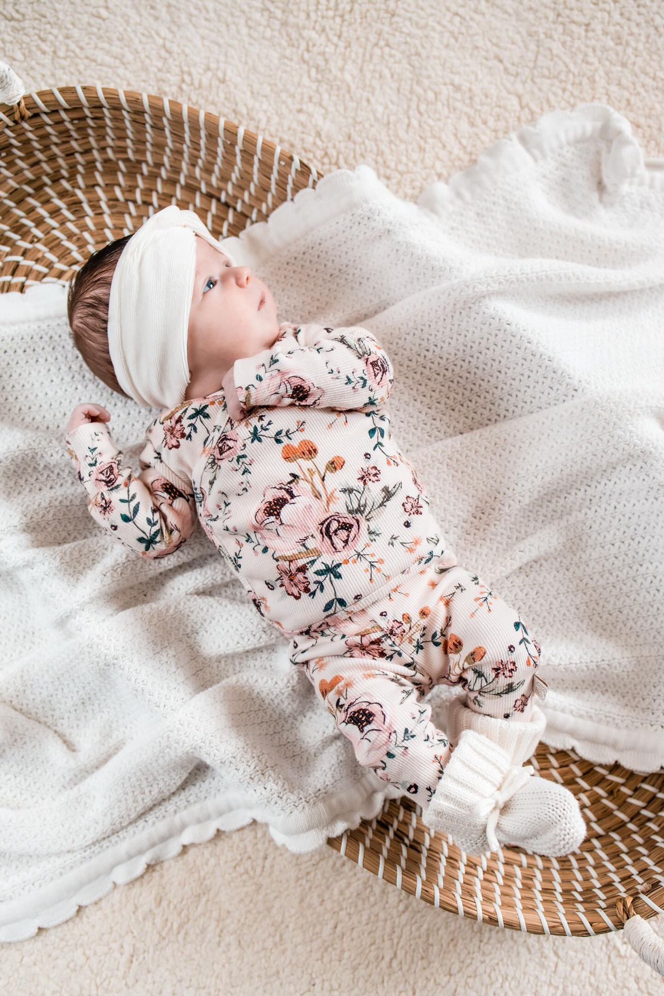 Een serene baby, gekleed in een gebloemd OEKO TEX-gecertificeerd katoenen Broekje Blamy-onesie van Yumi Baby en een witte hoofdband, ligt comfortabel op een gebreide deken in een geweven mand en staart verwonderd omhoog.