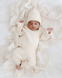 Een vredige pasgeboren baby, gekleed in witte kleding, lag comfortabel in een klein rotan wiegje, met een zachte deken en een delicate Yumi Baby Knitted Muts Pearl Whisper 0-3 mnd zachtjes gedrapeerd over de zijkant.