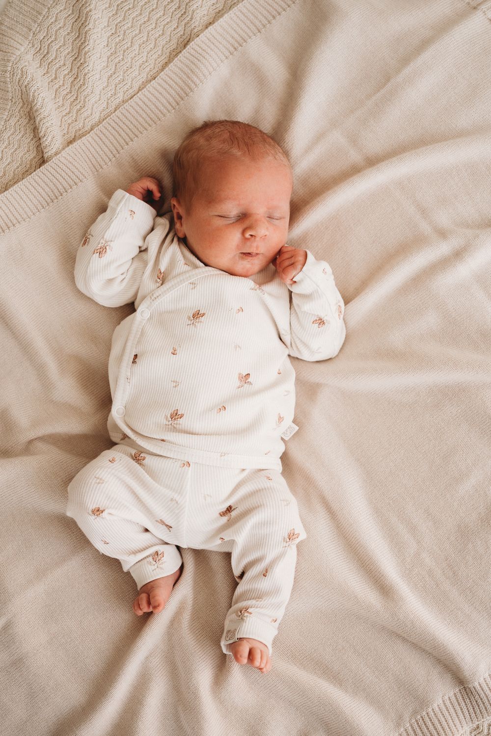 Een vredige pasgeboren baby, gekleed in een witte outfit van OEKO TEX-katoen met kleine printjes, heerlijk slapend op een beige deken met textuur.